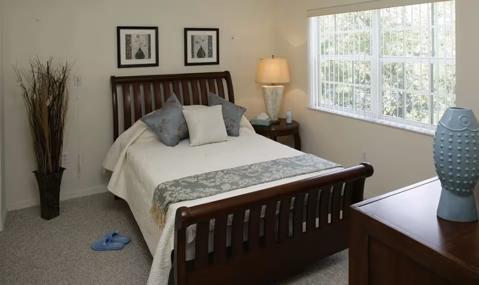 A cozy bedroom with a wooden bed frame, white bedding, and decorative pillows. There is a bedside table with a lamp and tissues, a large window with vertical blinds letting in natural light, a tall vase with dried branches in the corner, and a wooden dresser with a blue decorative vase on top. Two framed artworks hang on the wall above the bed.