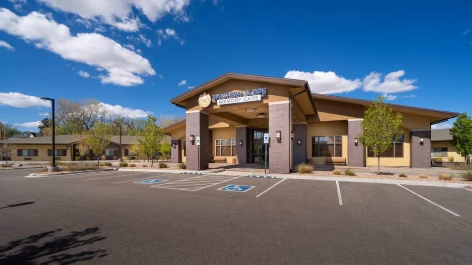 Front exterior of the Western Slope Memory Care building with entrance, benches and a nearly empty parking lot with accessible spaces under a blue sky.