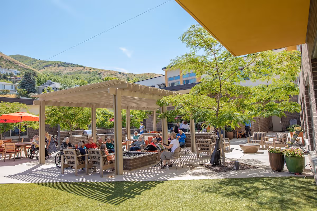 Outdoor patio area at The Ridge Foothill with elderly residents sitting under a pergola and around tables, enjoying the sunny day. There are trees providing shade, potted plants, and a backdrop of hills and residential houses.
