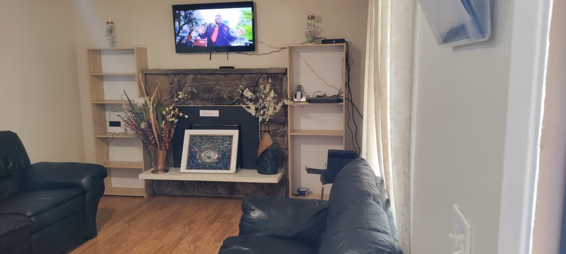 Living room with leather sofas, a wall-mounted TV above a stone fireplace, shelving units, decorative vases, and hardwood floor.