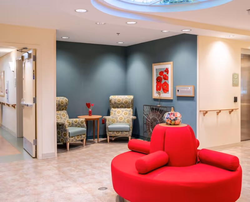 A cozy seating area in a senior living facility with two patterned armchairs and a small wooden table against a teal accent wall. A framed picture of red flowers hangs on the wall. In the foreground, there is a bright red circular cushioned bench with cylindrical armrests and a decorative vase on top. The area is well-lit with recessed ceiling lights and has a light-colored floor. A hallway and an elevator are visible in the background.