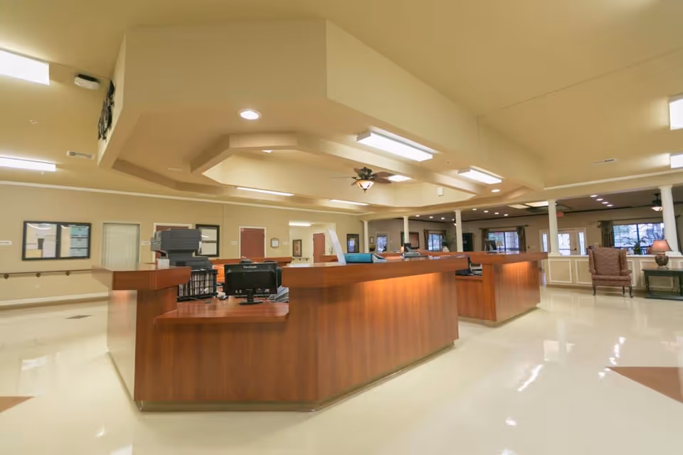 A spacious reception area with a large wooden front desk in the center. The room has beige walls and ceiling with recessed lighting and ceiling fans. There are chairs and tables along the walls and windows in the background letting in natural light.
