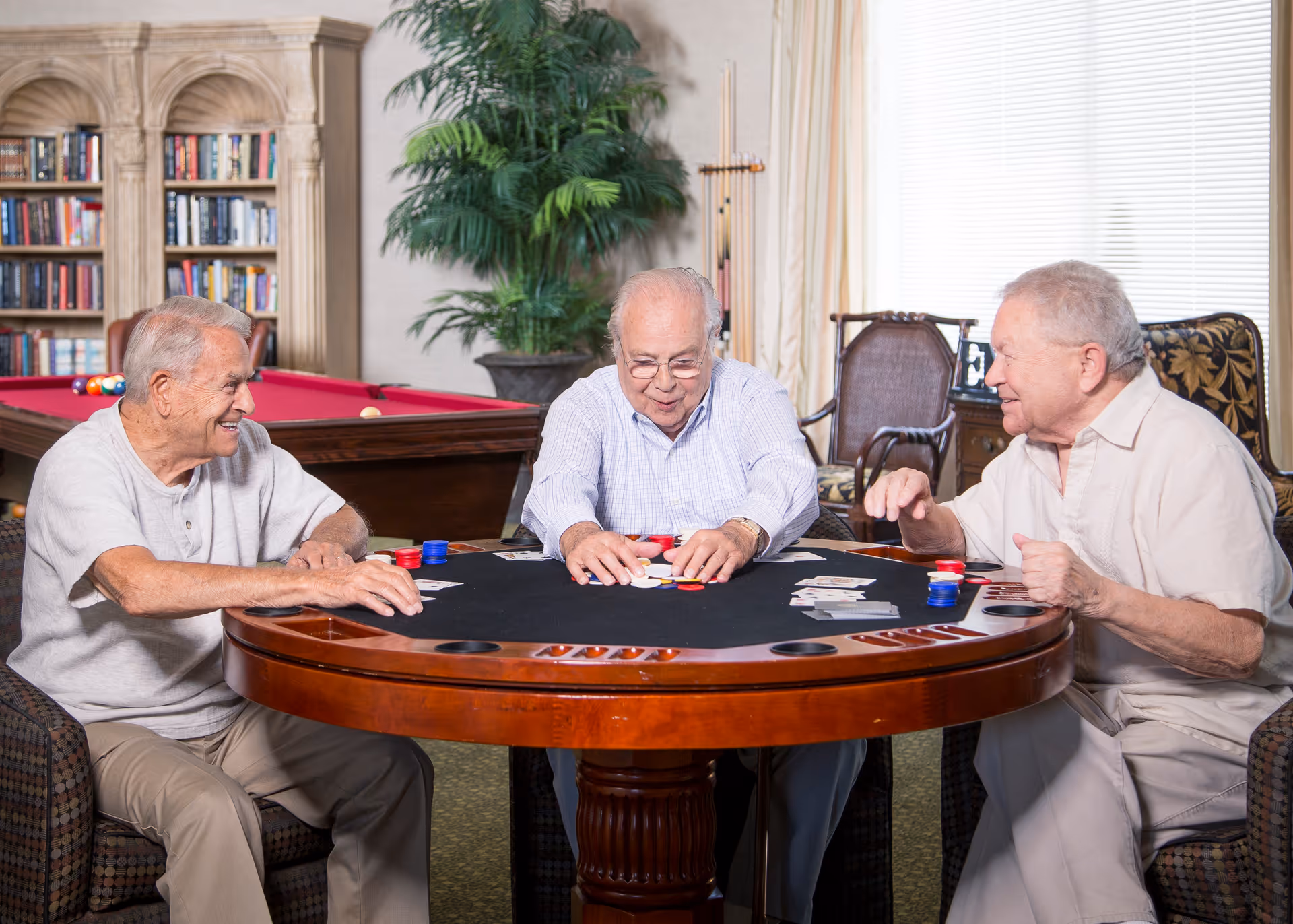 Three elderly men sitting around a round poker table playing cards and poker chips in a cozy room with a pool table, bookshelf, and large plant in the background.