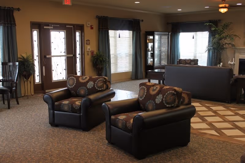 A cozy living room area in a senior living facility with two patterned armchairs in the foreground, a dark sofa in the background, large windows with curtains, a glass-front cabinet, potted plants, and a ceiling fan. The room has a mix of carpet and tiled flooring and a double glass door entrance.