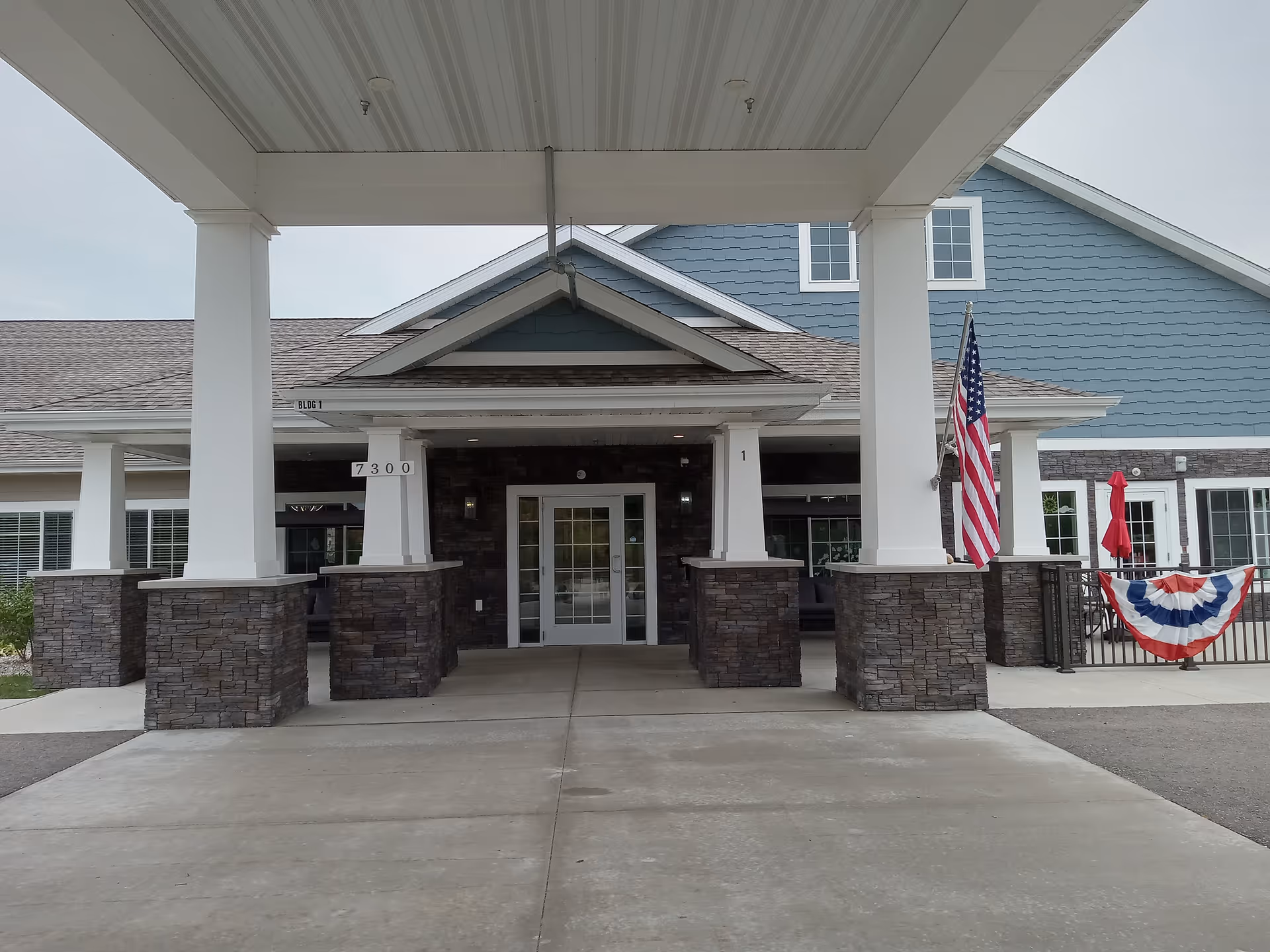 Covered front entrance of a senior living building with stone pillars, glass double doors, and an American flag.