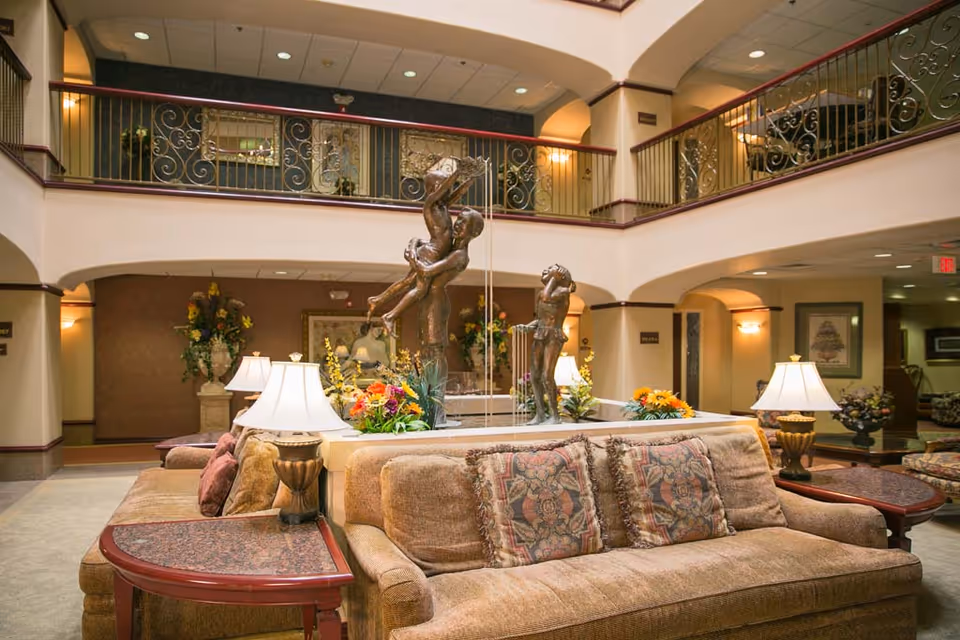 A spacious and elegant two-story lobby area with a central bronze sculpture of children playing, surrounded by floral arrangements. Comfortable beige sofas with patterned cushions and side tables with lamps are arranged around the sculpture. The upper level features decorative wrought iron railings overlooking the lobby.