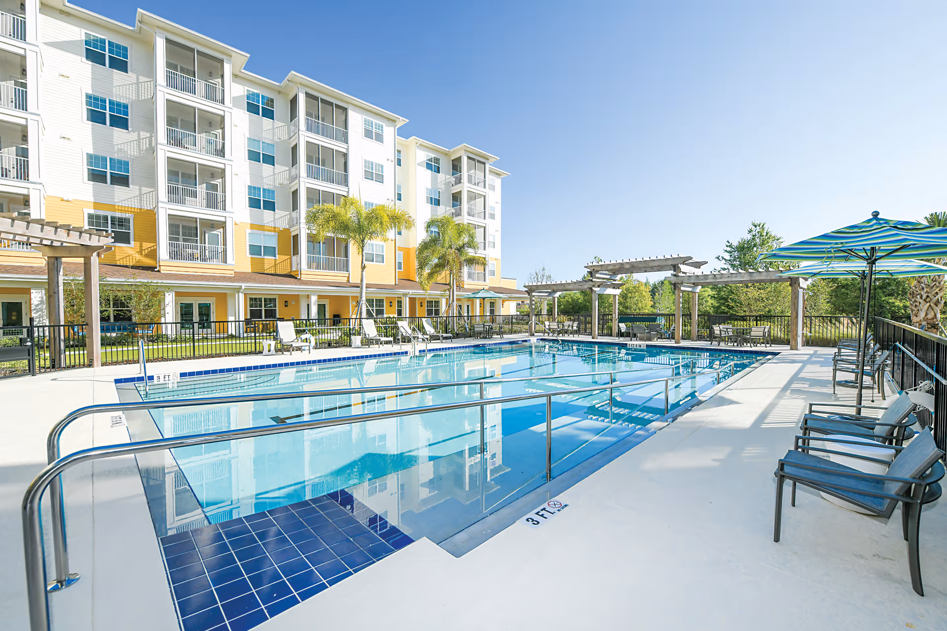 Outdoor swimming pool area at Discovery Village Trinity with clear blue water, surrounded by lounge chairs, tables with umbrellas, palm trees, and a multi-story residential building in the background under a clear blue sky.
