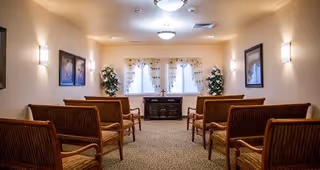 A small chapel or meditation room with wooden pew-style seating arranged in two rows facing a window with sheer curtains. There are two framed pictures on the walls, two potted plants near the window, and soft lighting from ceiling fixtures and wall sconces.