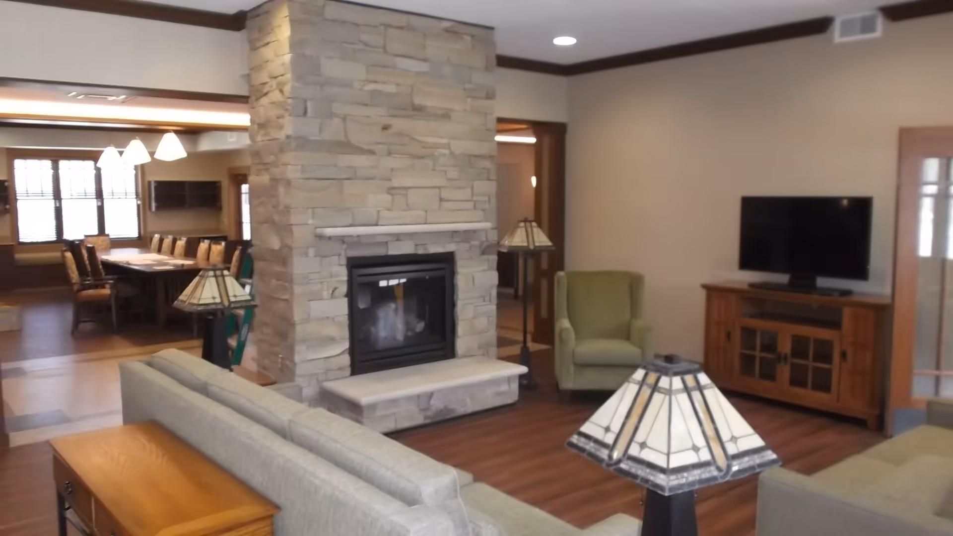 A cozy living room area featuring a stone fireplace in the center, surrounded by comfortable seating including a green armchair and gray sofas. A wooden TV stand with a flat-screen television is positioned against the wall. In the background, a dining area with a long table and chairs is visible, illuminated by hanging lights.