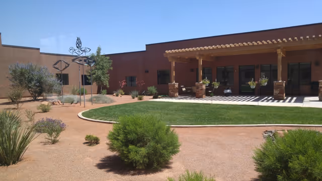 Outdoor courtyard area at Southern Utah Veterans Home – Ivins featuring a green lawn surrounded by desert landscaping with bushes and small trees, a covered patio with seating, and a clear blue sky.