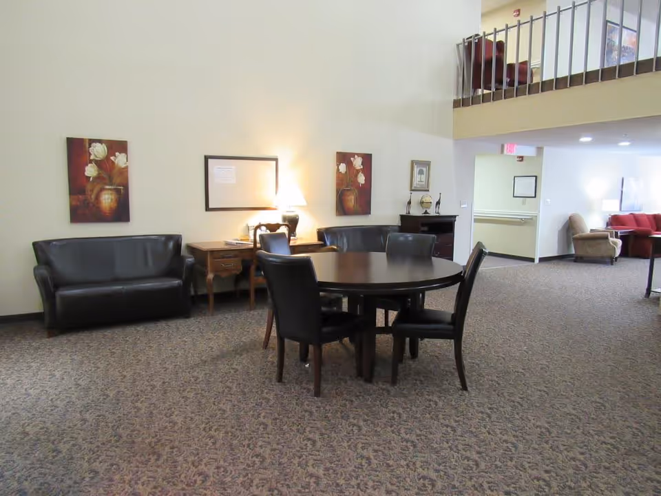 Spacious senior living common area with a round table and chairs, sofas, artwork on the walls, and a lofted railing.