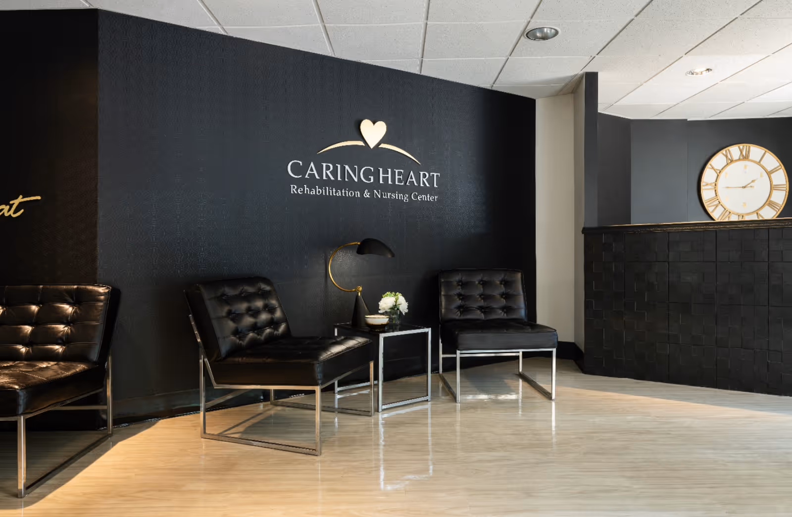Reception area of Caring Heart Rehabilitation and Nursing Center with black leather chairs, a small table with a lamp and flowers, a black textured wall with the facility's name and logo, and a large clock on a dark wall behind the reception desk.