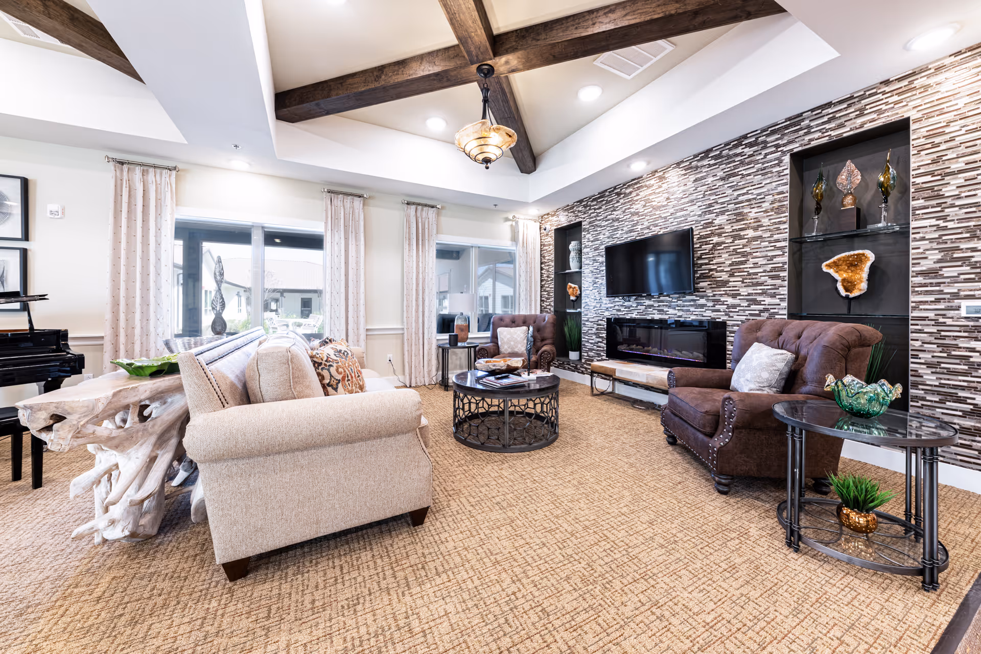 A bright and spacious living room with beige carpet and a high ceiling featuring exposed wooden beams. The room includes a beige sofa, two brown armchairs with pillows, a round coffee table, and side tables with decorative items. A modern electric fireplace is set into a tiled accent wall beneath a mounted flat-screen TV. Large windows with light curtains allow natural light to fill the space, and a black grand piano is visible in the corner.