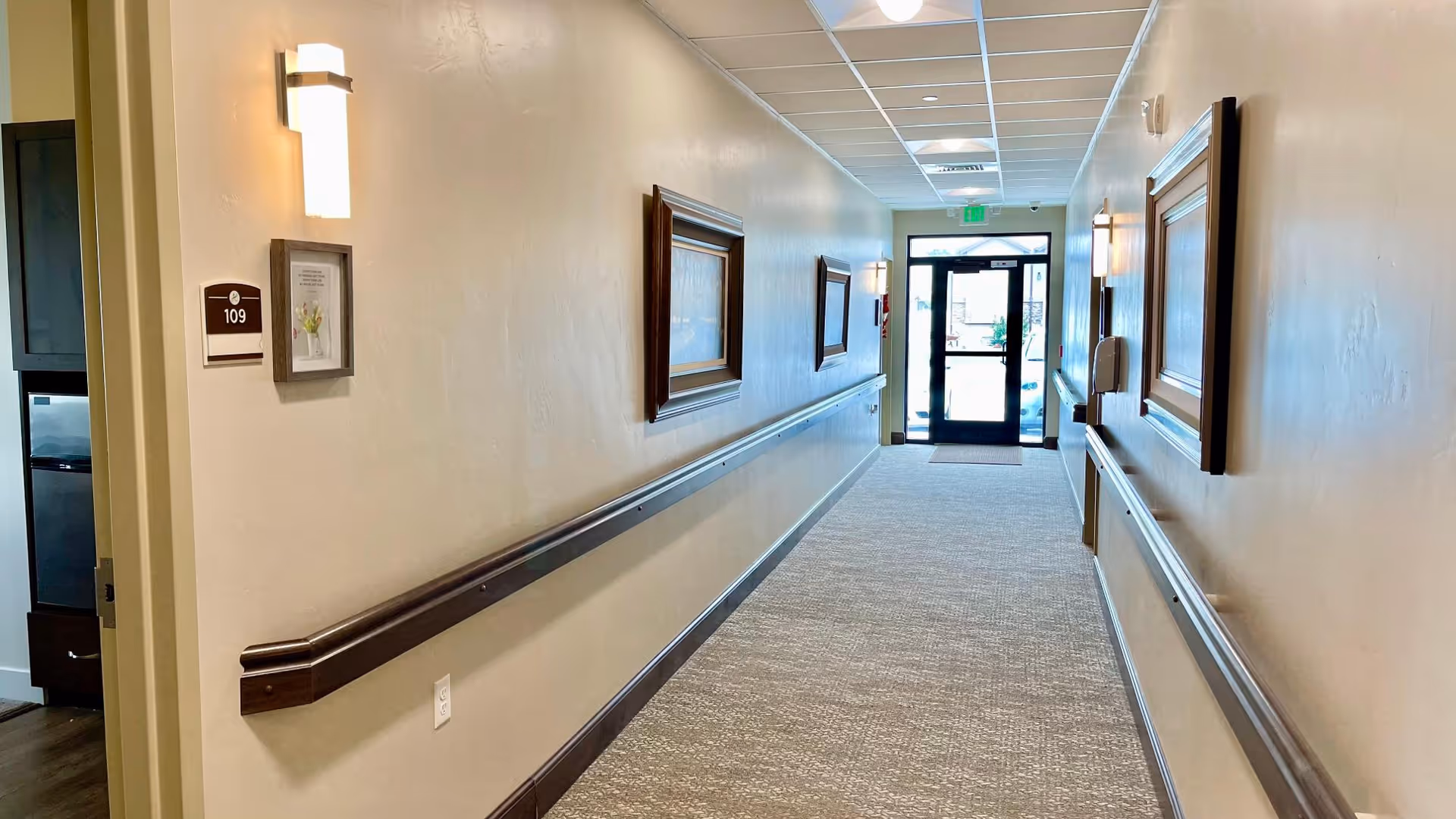 A well-lit hallway in an assisted living facility with beige walls, carpeted floor, and wooden handrails on both sides. There are framed pictures on the walls and a door labeled 109 on the left. The hallway leads to a glass exit door with natural light coming through.
