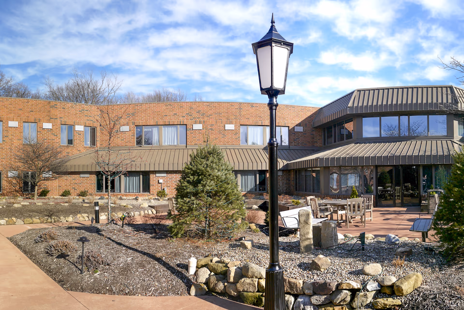 Outdoor courtyard area of Pleasant Lake Villa with a brick building in the background, a black lamppost in the center, landscaped garden beds with rocks and small trees, and patio seating with tables and chairs under a partly cloudy sky.