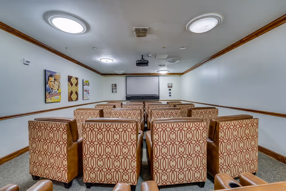 A small theater room with patterned upholstered chairs arranged in rows facing a large projection screen on the front wall. The room has white walls with wooden trim, ceiling lights, and movie posters on the left wall.
