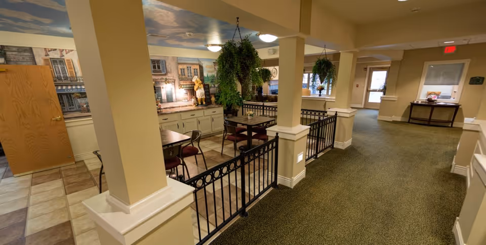 Interior common area with tables and chairs, hanging plants, a muraled wall and a carpeted corridor in a senior living facility.