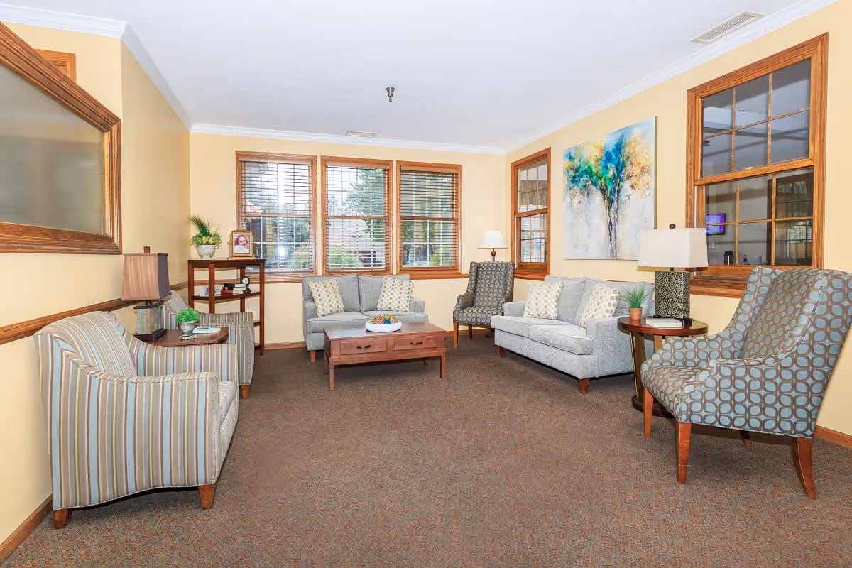 A cozy living room with beige walls and brown carpet, featuring two gray sofas with patterned cushions, two patterned armchairs, a wooden coffee table with a decorative bowl, a small wooden shelf with a framed photo and plants, two table lamps, and a colorful abstract tree painting on the wall. Large windows with wooden blinds allow natural light into the room.