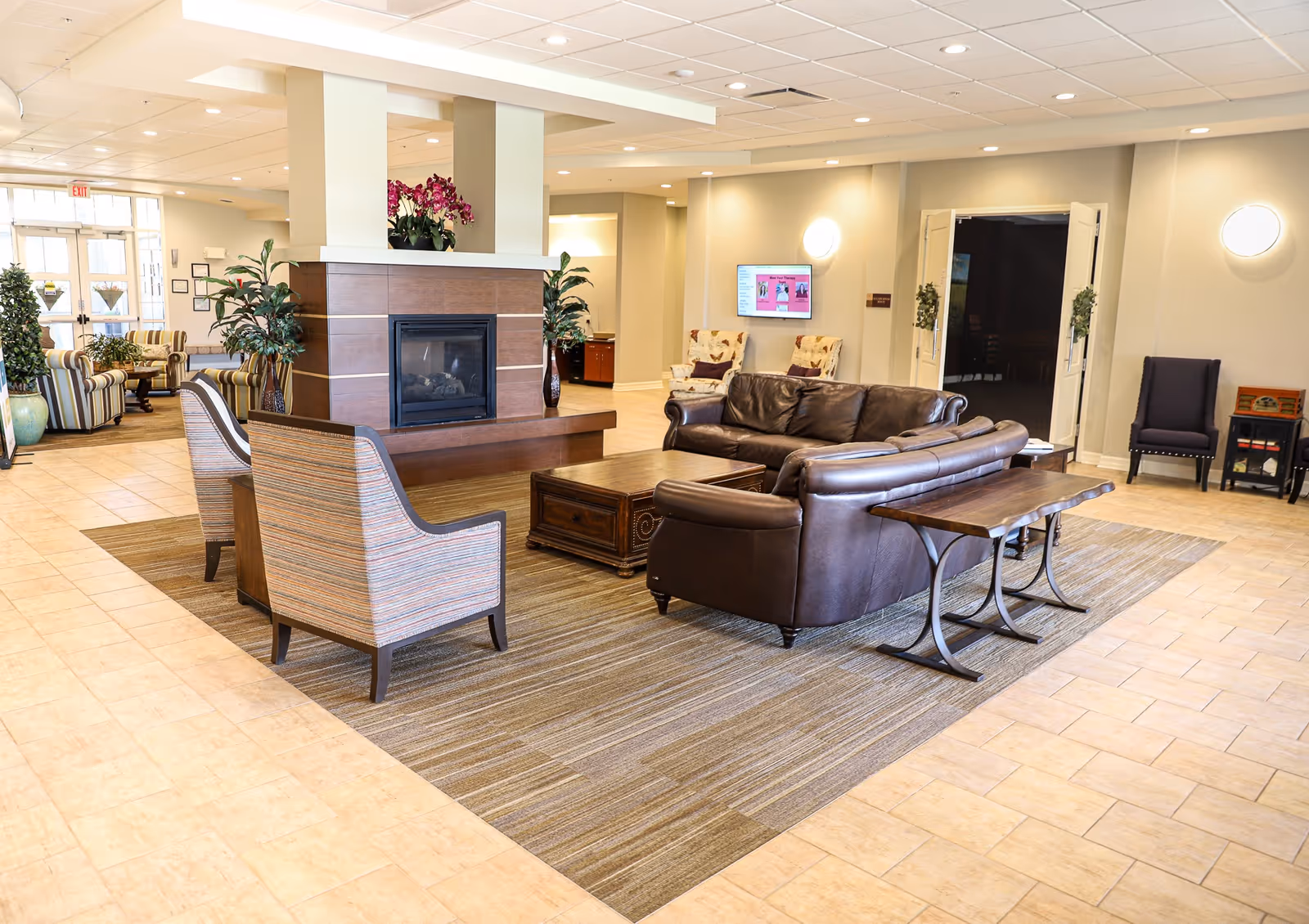 A spacious senior living facility common area with a central fireplace surrounded by two striped armchairs, a brown leather sofa, and a wooden coffee table on a large area rug. The room has tiled floors, potted plants, and additional seating including two floral armchairs near a wall-mounted TV. Double doors and wall sconces are visible in the background.