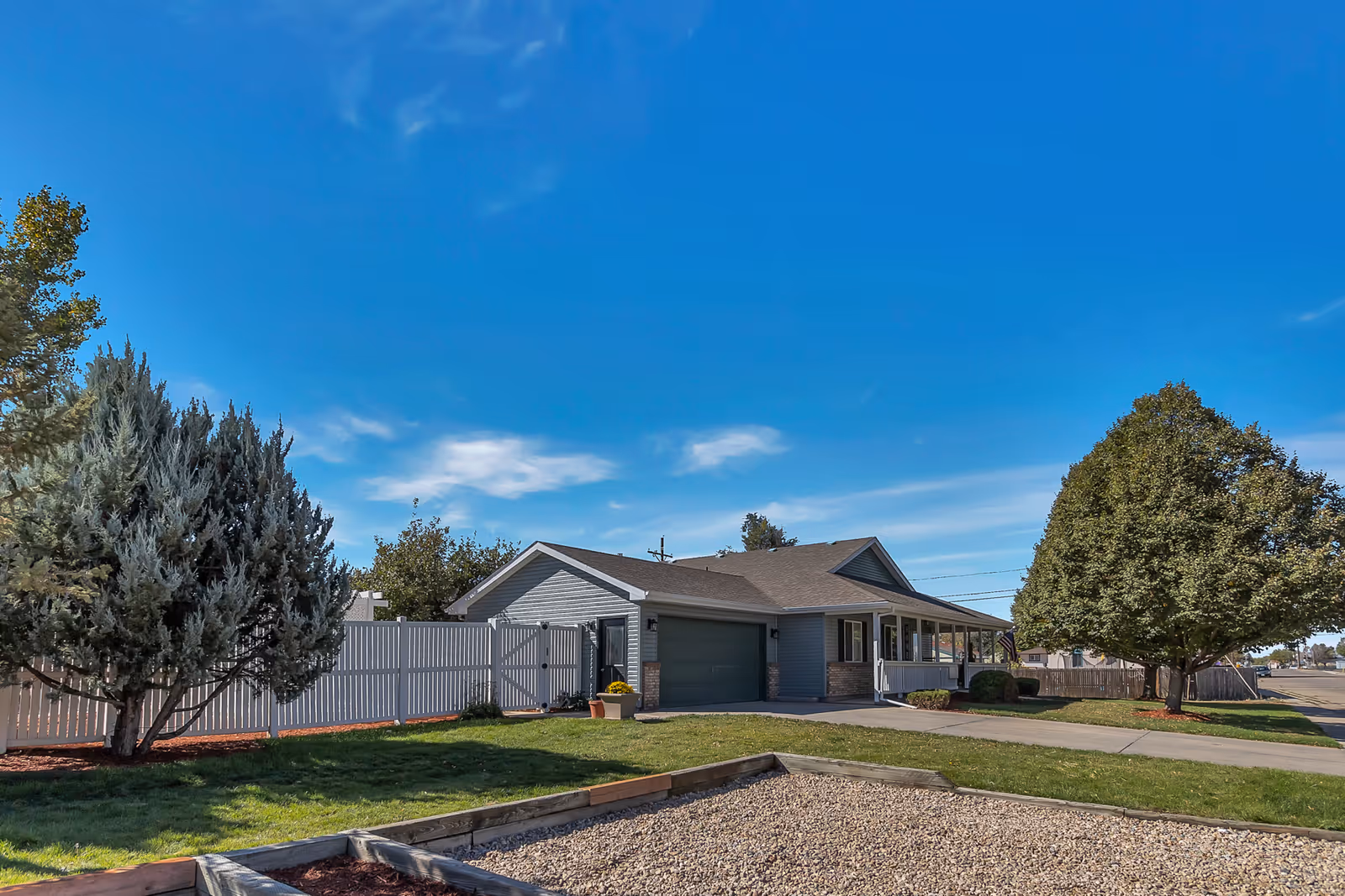 Exterior view of a single-story residential building with a garage, surrounded by a white fence, green lawn, trees, and a clear blue sky.