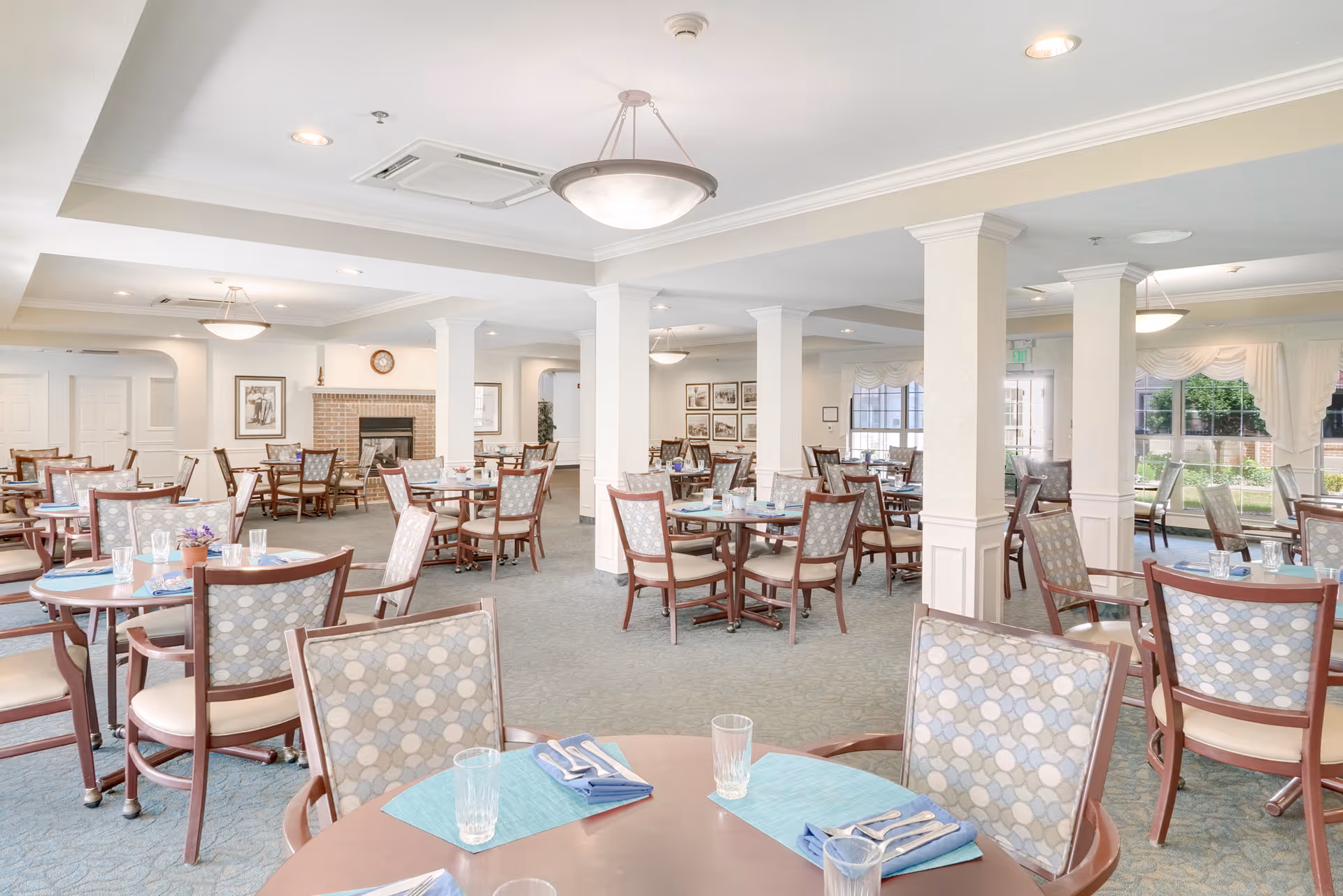 A spacious dining room in a senior living facility with multiple round tables set with placemats, napkins, utensils, and glasses. The room features patterned chairs, carpeted floors, white walls with columns, ceiling lights, and large windows allowing natural light. A fireplace and framed pictures are visible in the background.