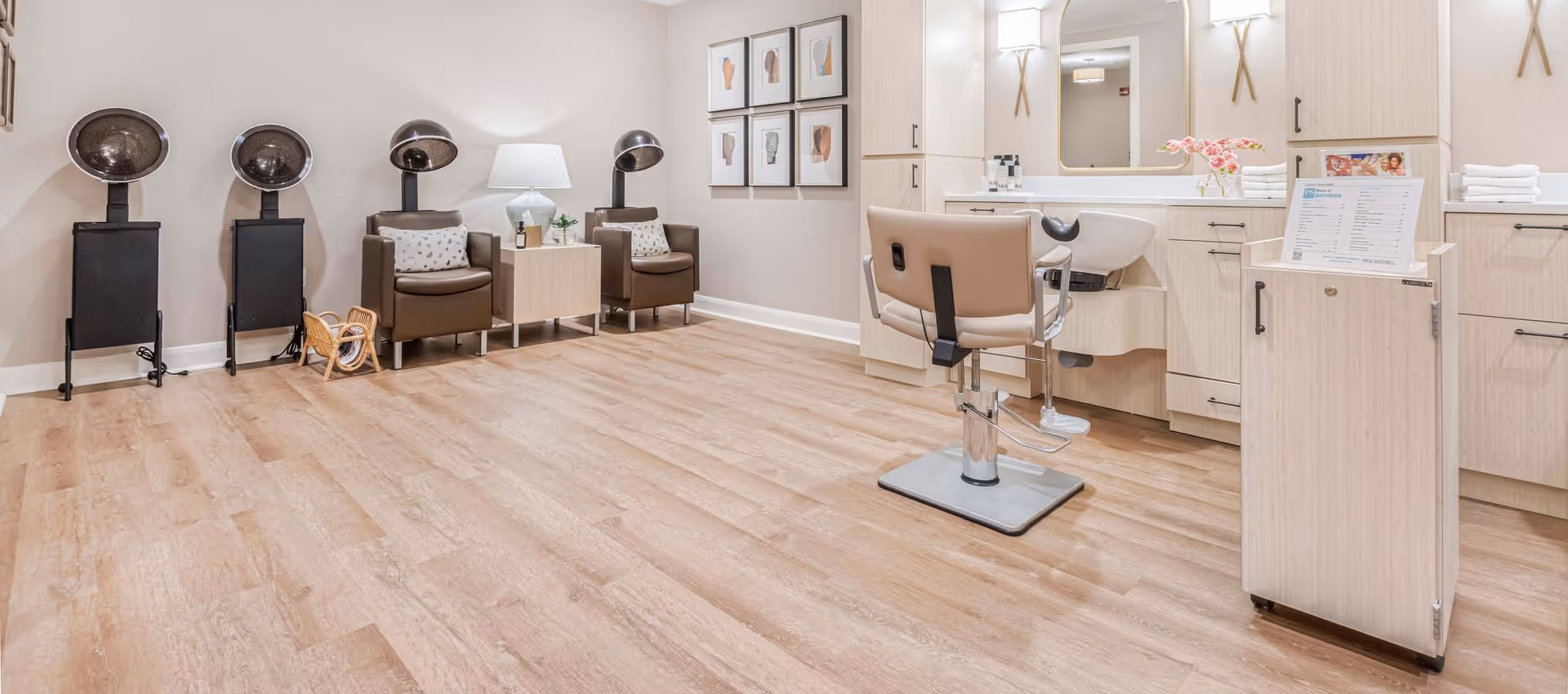 Interior of a salon area in a senior living facility with three hair drying stations, two brown leather chairs with pillows, a small table with a lamp and plant, a salon chair in front of a sink and mirror, light wood cabinetry, and framed artwork on the wall.