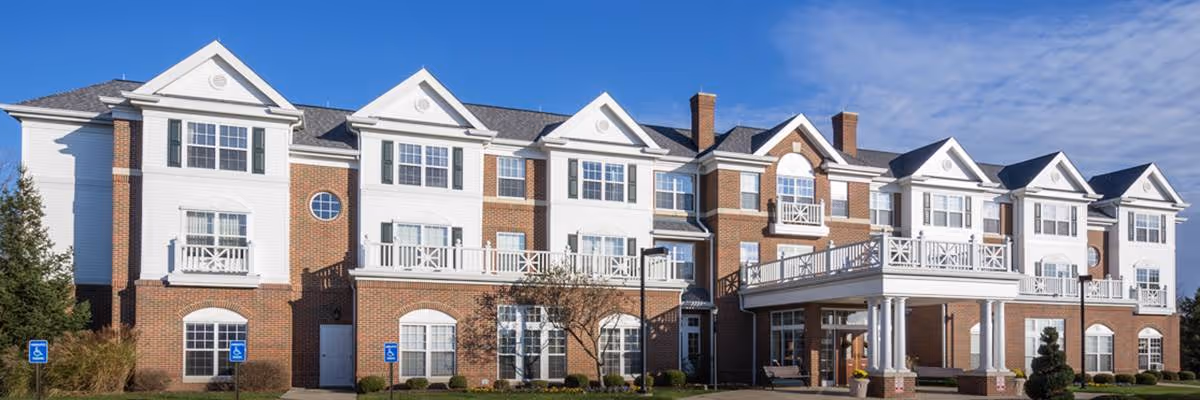Front exterior of a three-story brick-and-white senior living building with balconies, an entrance portico, and landscaped grounds under a blue sky.