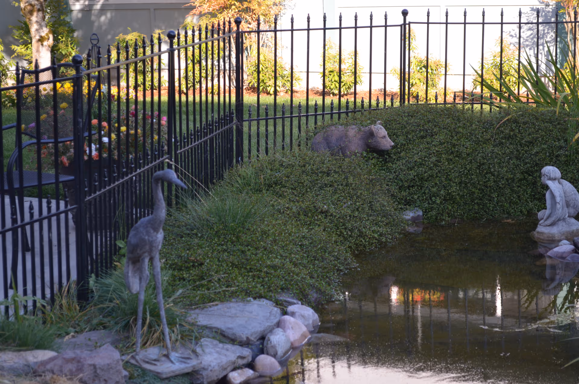 A fenced garden area with a small pond surrounded by rocks and greenery. There are three statues near the pond: a bird, a bear, and a seated figure. In the background, there are plants and flowers with a gray fence and building wall.