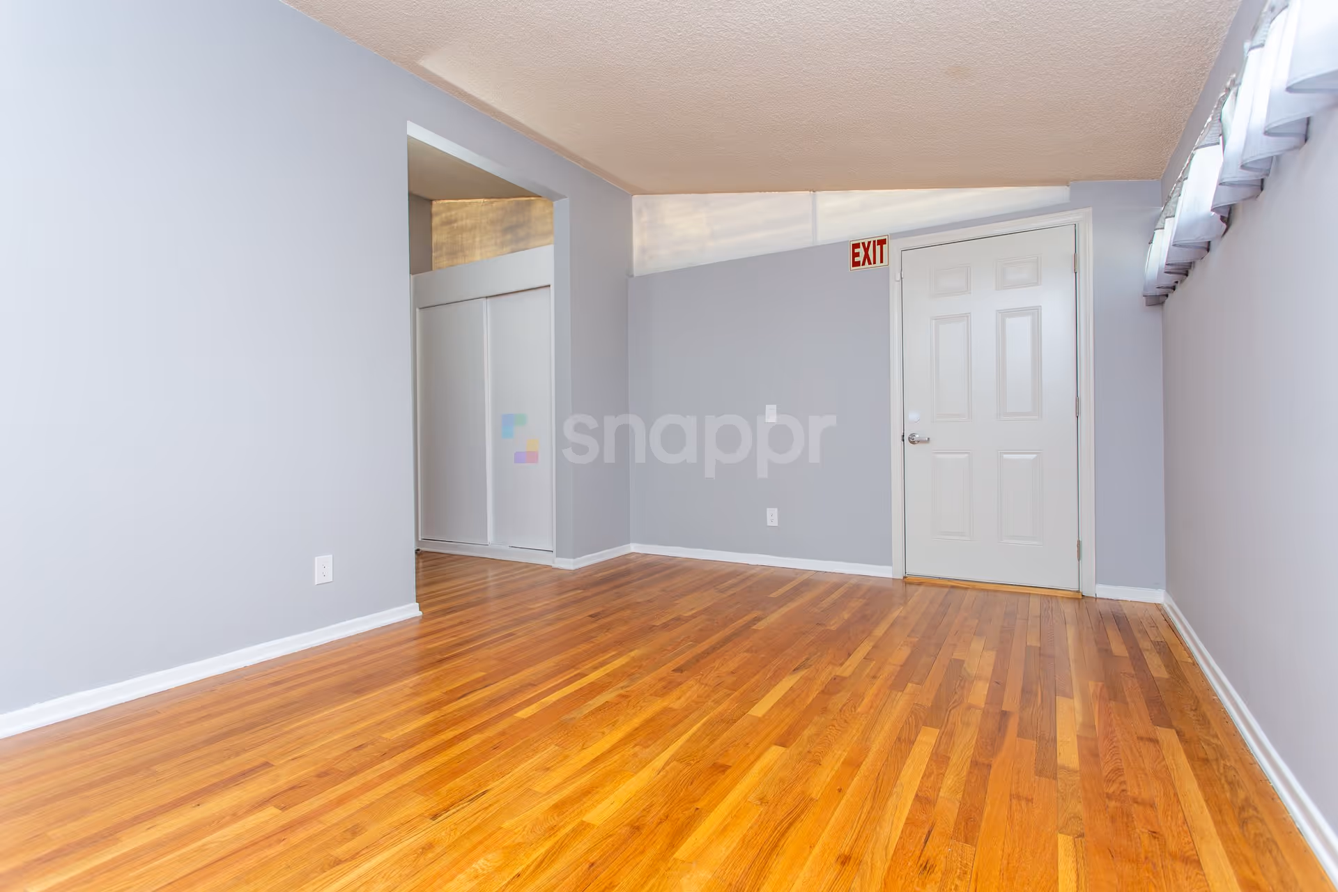 Empty room with light gray walls, wooden floor, a white door with an exit sign above it, and a closet with sliding doors. A row of windows with white curtains is visible on the right side.