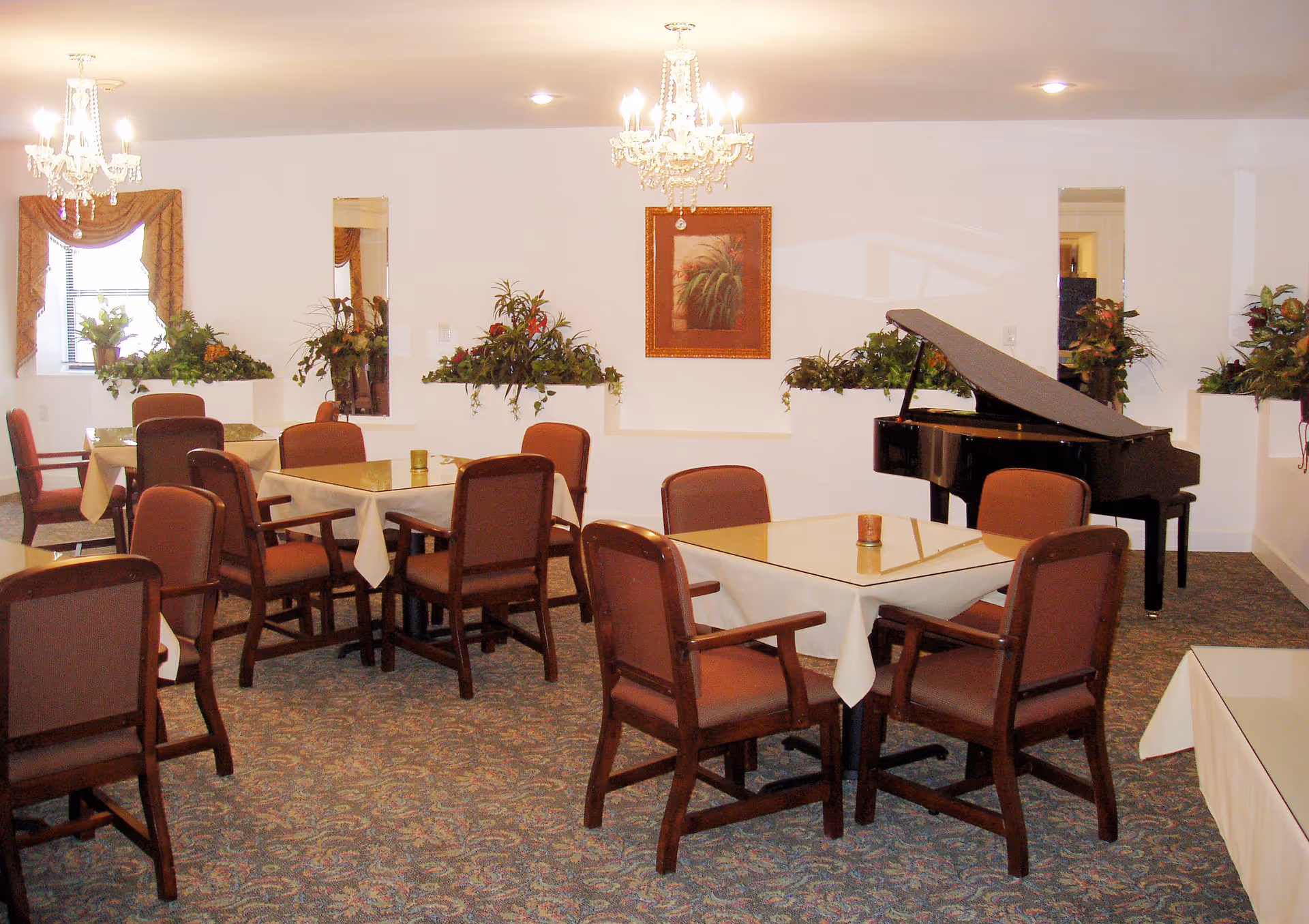 A dining room with several tables covered with white tablecloths and surrounded by brown cushioned chairs. The room features a grand piano on the right side, decorative plants along the walls, two chandeliers hanging from the ceiling, and a framed picture on the wall. The carpet has a patterned design, and there are windows with curtains on the left side.
