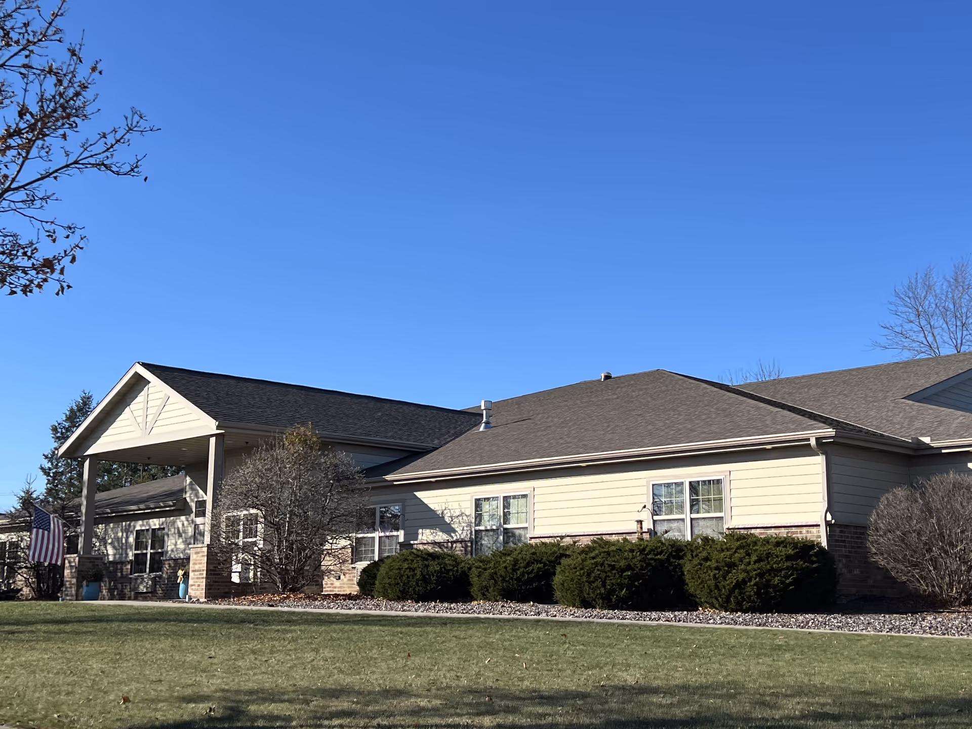 Exterior view of a single-story senior living facility building with beige siding, a dark shingled roof, and several windows. The building is surrounded by neatly trimmed bushes and a well-maintained lawn under a clear blue sky. An American flag is visible near the entrance.