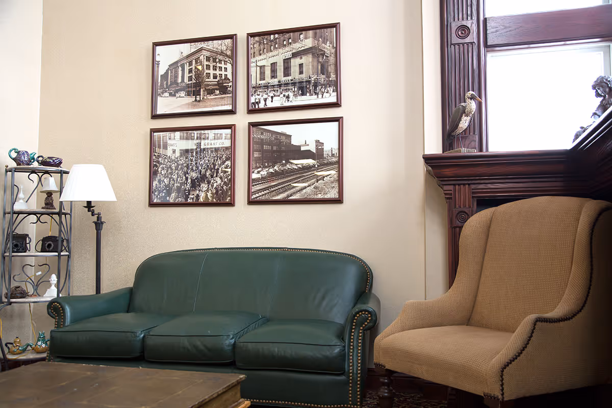 A cozy sitting area with a green leather couch and a beige upholstered armchair. On the wall above the couch are four framed black and white vintage photographs. To the left is a metal shelving unit with decorative items and a floor lamp. To the right is a wooden window frame with a decorative bird sculpture on the windowsill.