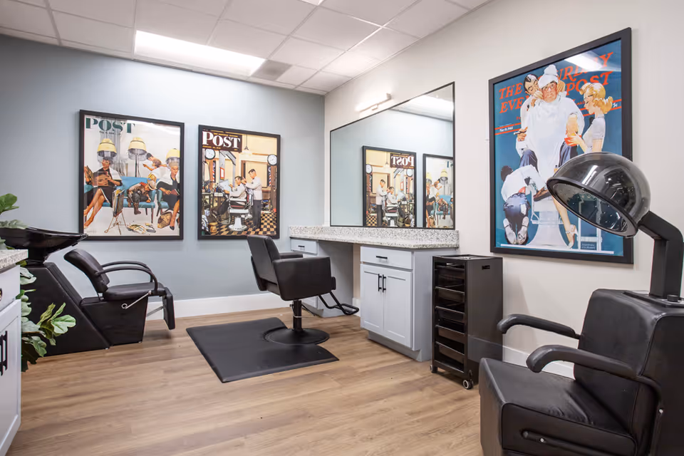 Interior salon room with a styling chair, hooded dryer, shampoo sinks, large mirror and framed vintage posters.