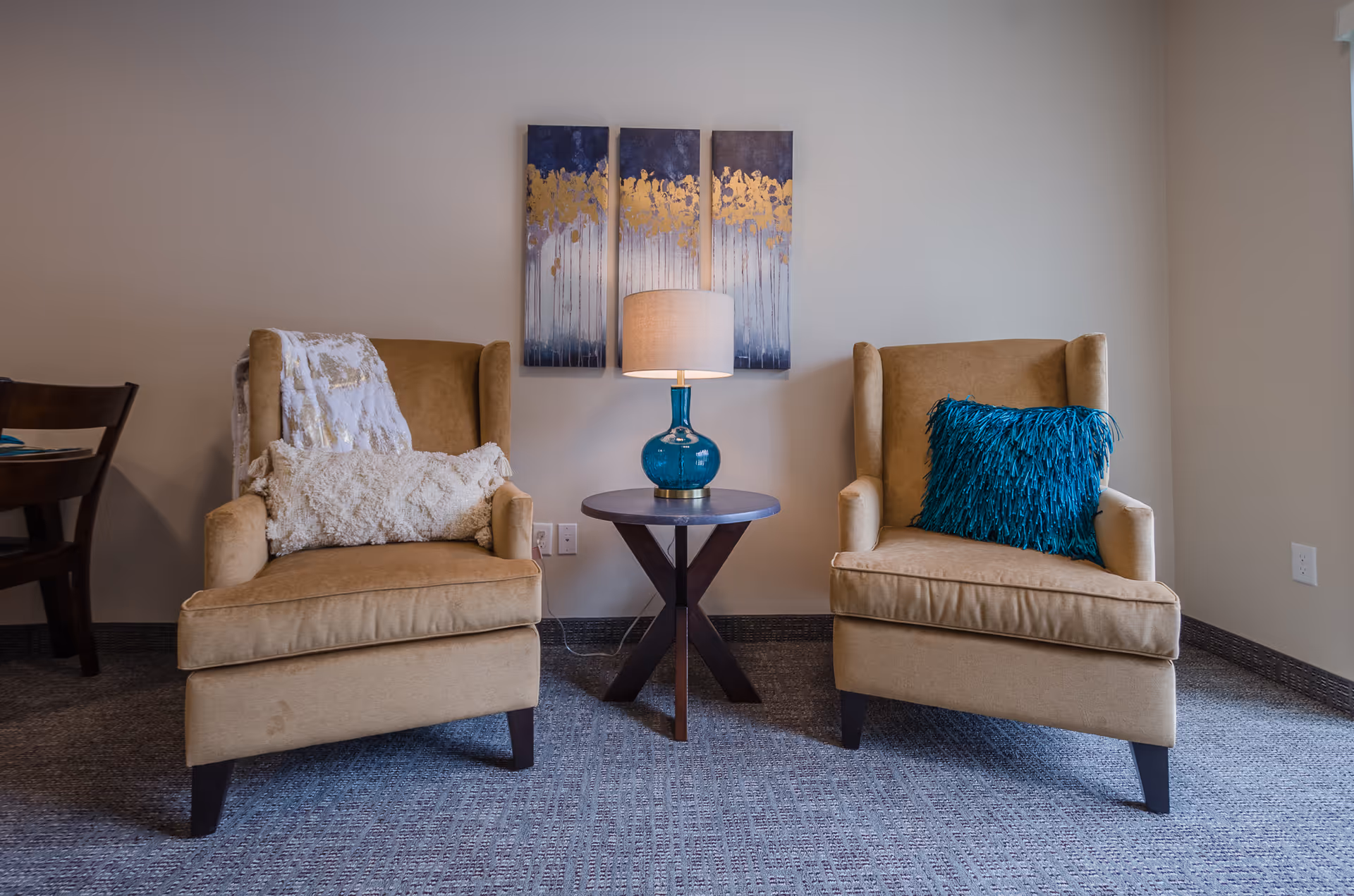 Two beige armchairs with decorative pillows and a throw blanket are positioned on either side of a small round wooden table with a blue glass lamp. Behind the table, three vertical abstract paintings with blue and gold tones hang on a light-colored wall. The floor is carpeted in a gray pattern.
