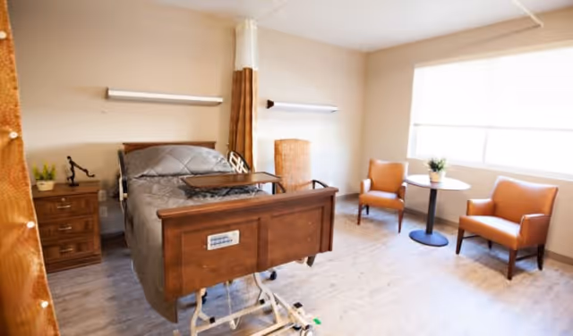 A bright and clean patient room with a hospital bed featuring a gray quilted blanket, a wooden nightstand with a small plant and lamp, two orange armchairs, and a round table with a small plant near a large window with blinds.