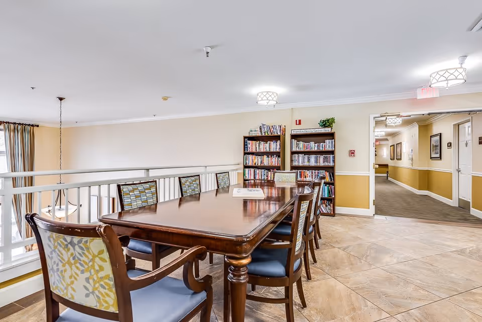 A well-lit interior common area in a senior living facility featuring a long wooden table surrounded by chairs with patterned upholstery. Behind the table are two bookshelves filled with books and a small plant on top. The area overlooks a lower floor through white railings, and there is a hallway with framed pictures and doors visible in the background.