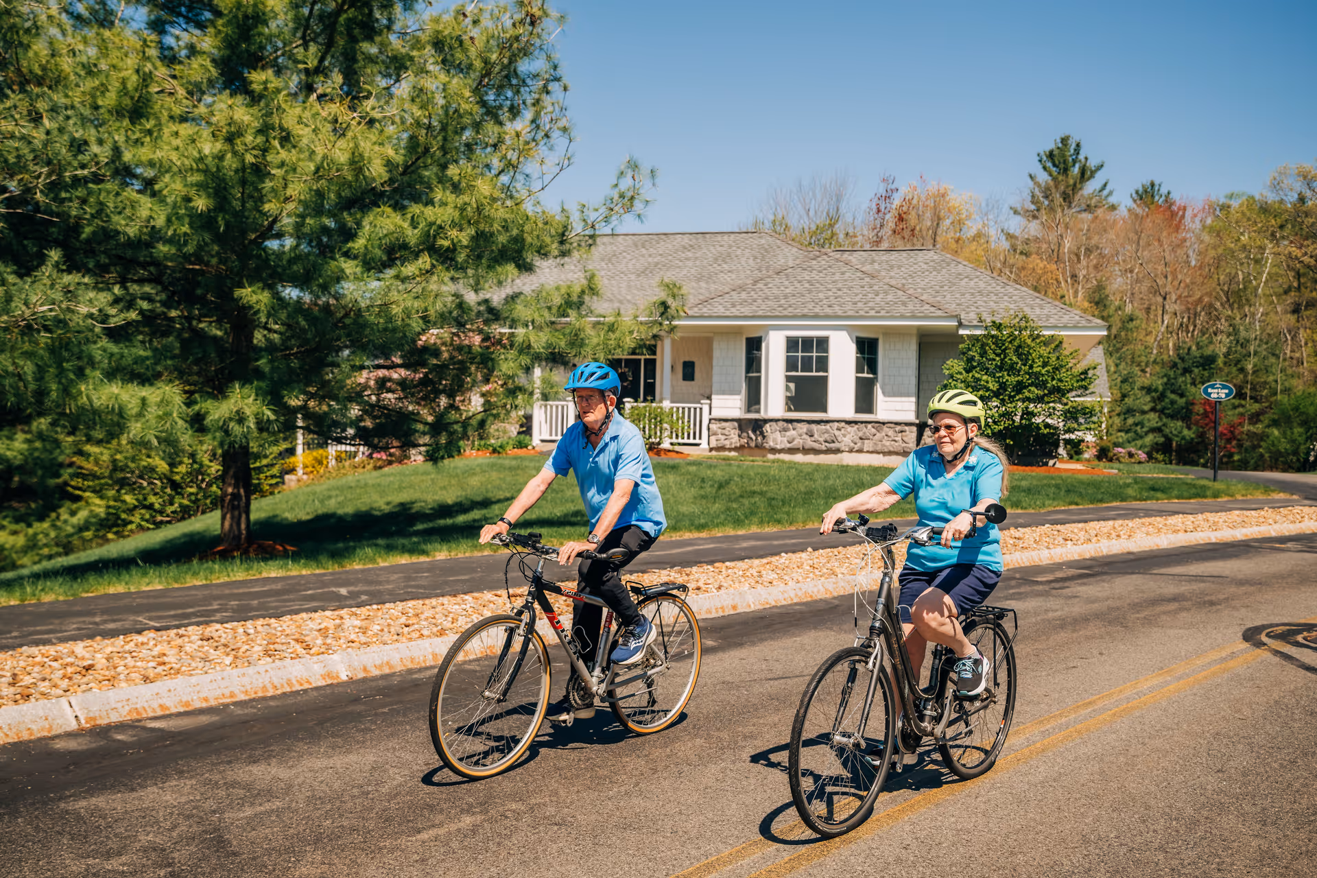 Two elderly people wearing helmets riding bicycles on a paved road in front of a single-story house surrounded by trees and greenery on a sunny day.