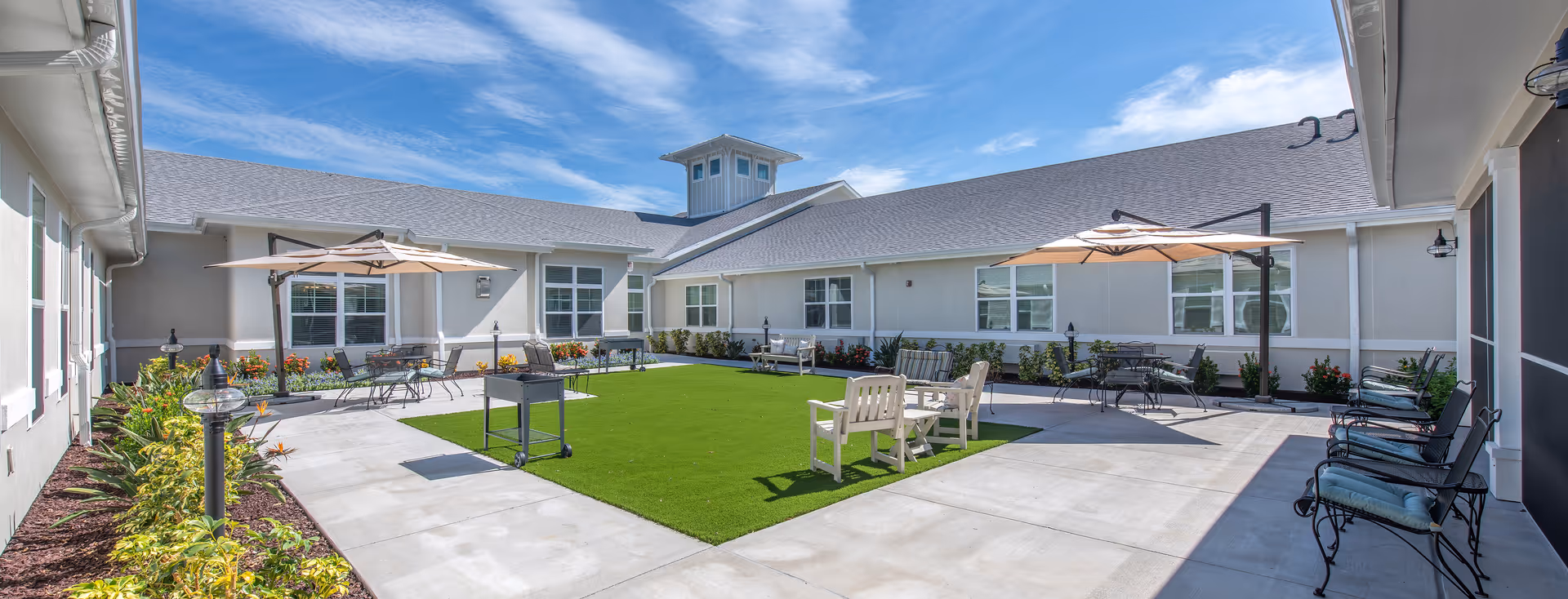 Outdoor courtyard area at The Addison of Narcoossee with green artificial turf in the center, surrounded by concrete walkways, patio tables with umbrellas, chairs, benches, and landscaping with flowers and shrubs under a blue sky with some clouds.