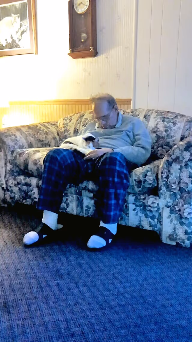 A person in plaid pajama pants sits on a floral couch in a living room holding a small black-and-white cat.