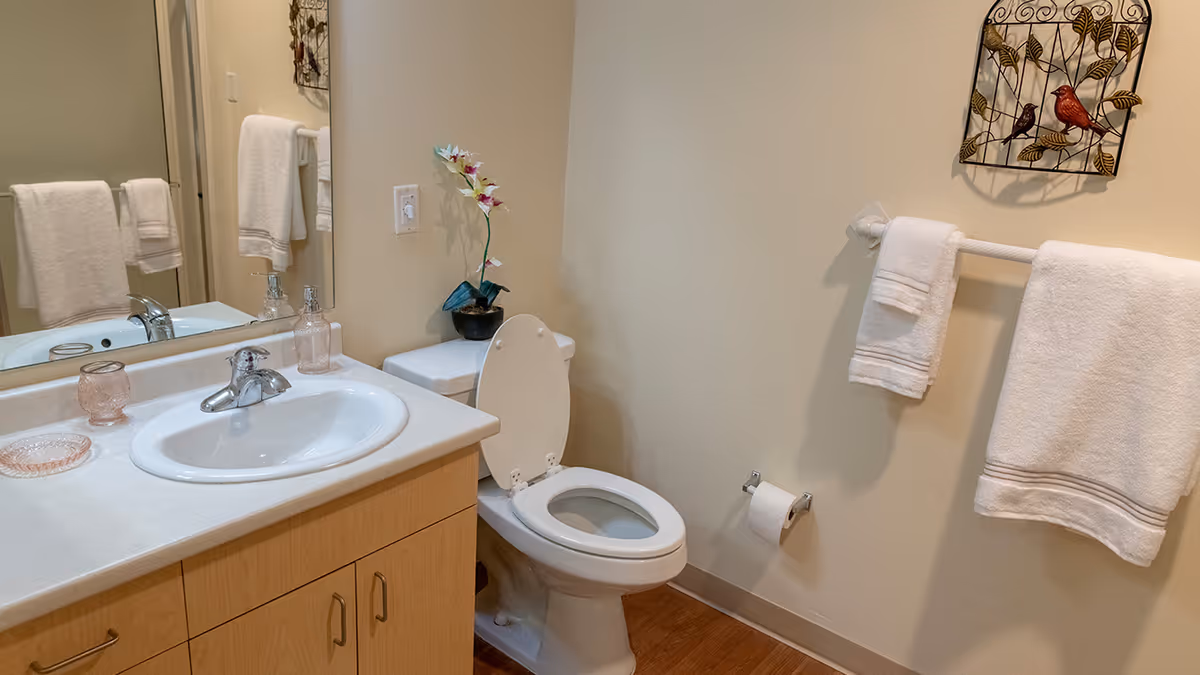 A clean bathroom with a white sink and countertop on a wooden cabinet, a toilet with the lid open, a small potted plant with flowers on the toilet tank, white towels hanging on a towel rack, a toilet paper holder with a roll of toilet paper, and a decorative wall hanging featuring birds on branches.