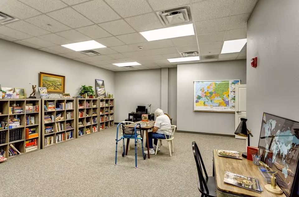 A communal interior room with bookshelves along one wall, tables and chairs, and a resident seated at a table.