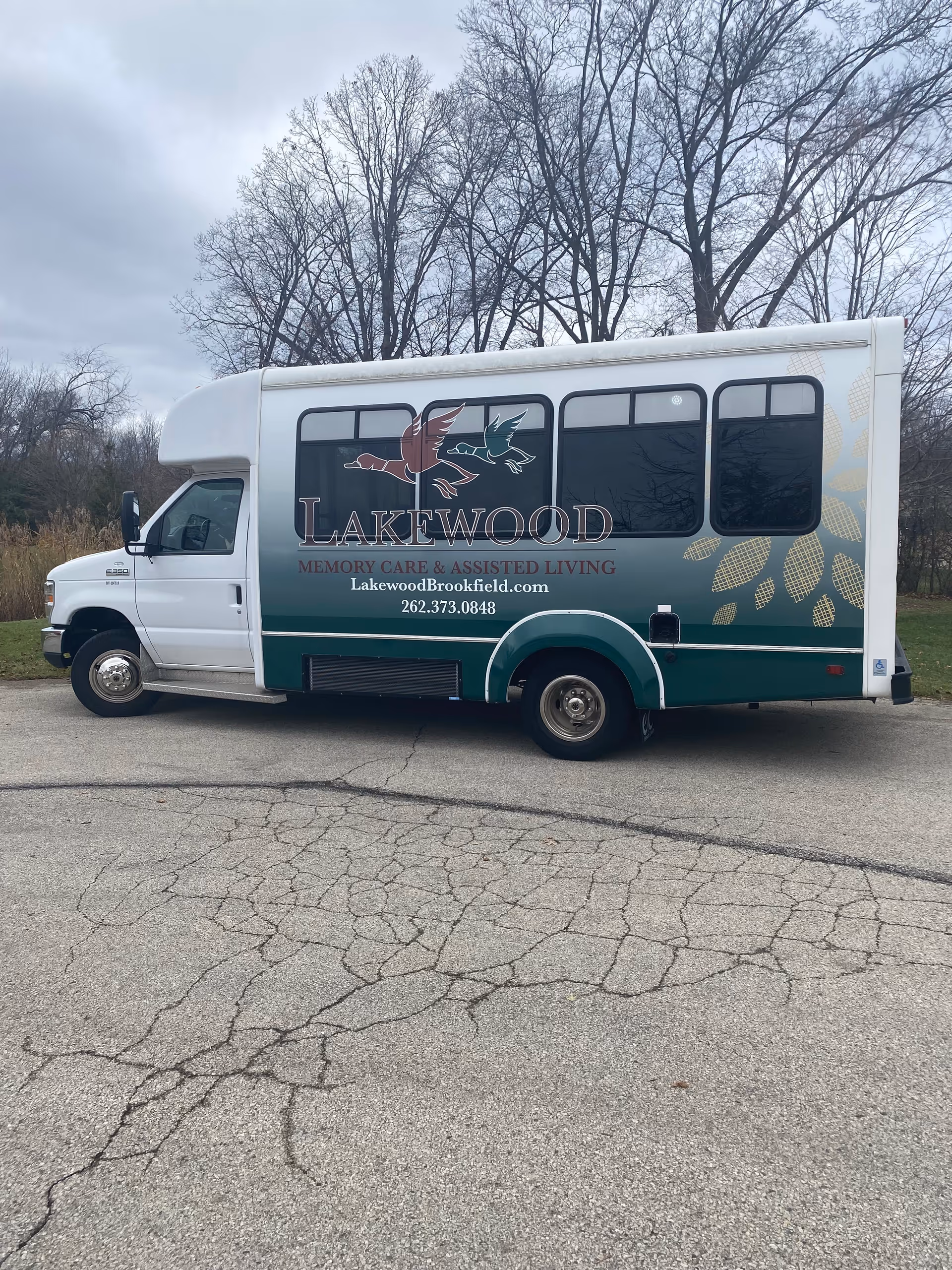 A Lakewood Memory Care & Assisted Living shuttle bus parked on cracked pavement with leafless trees in the background.
