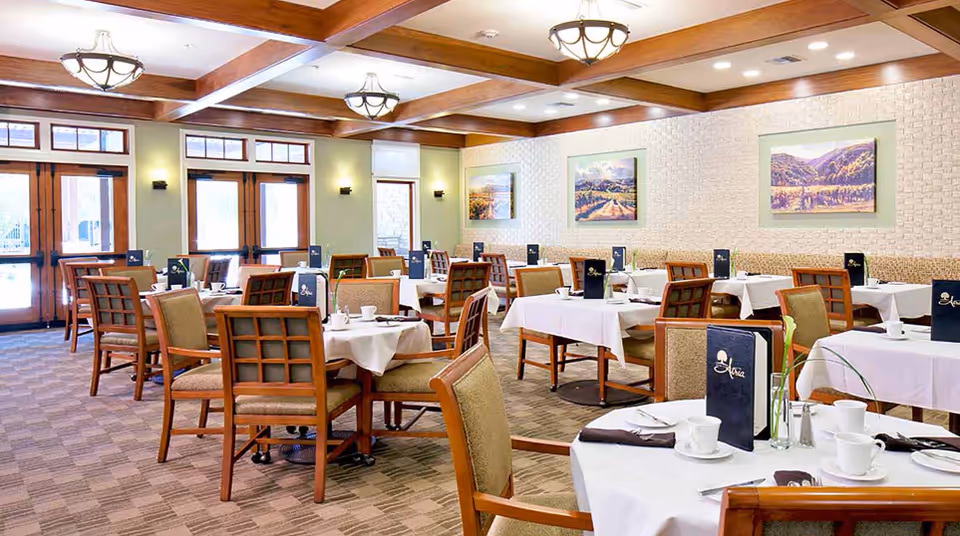 A bright and spacious dining room with multiple tables covered in white tablecloths, set with white cups, saucers, and silverware. Each table has a dark blue menu labeled 'Atria' and a small vase with a single flower. The room features wooden chairs with cushioned seats, large windows and doors letting in natural light, beige walls with framed landscape paintings, and a ceiling with wooden beams and decorative light fixtures.