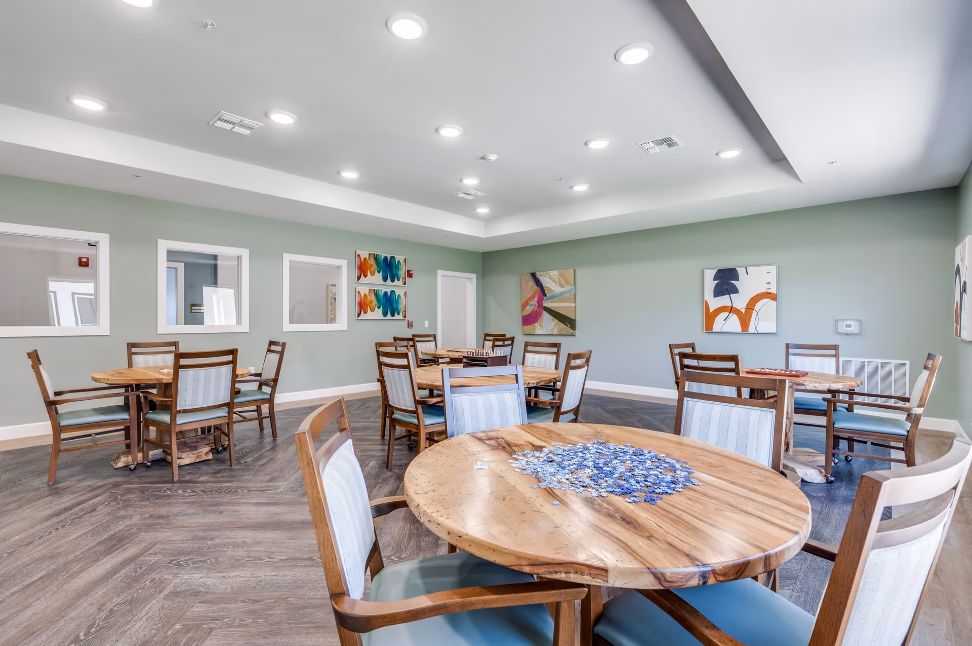 A spacious room with several round wooden tables and chairs arranged neatly. One table in the foreground has a partially completed blue and white jigsaw puzzle. The walls are painted light green and decorated with colorful abstract paintings. The ceiling has recessed lighting, and the floor has a wood pattern.