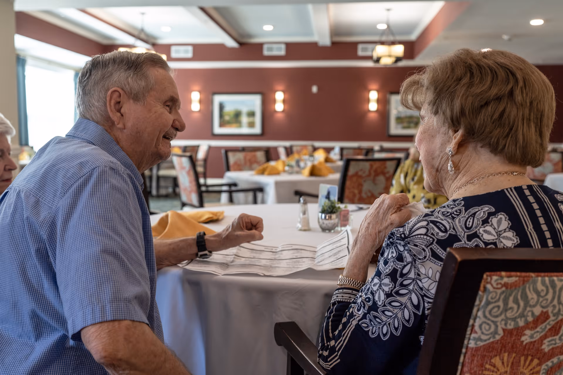 Two elderly people sitting at a table in a dining room, engaged in conversation. The table is set with a white tablecloth and yellow napkins. The room has red walls with framed pictures and wall sconces, and several other tables and chairs are visible in the background.