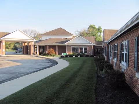 Exterior view of a senior living facility with brick buildings, a curved driveway, a covered entrance, and a well-maintained lawn with shrubs and trees in the background under a clear sky.