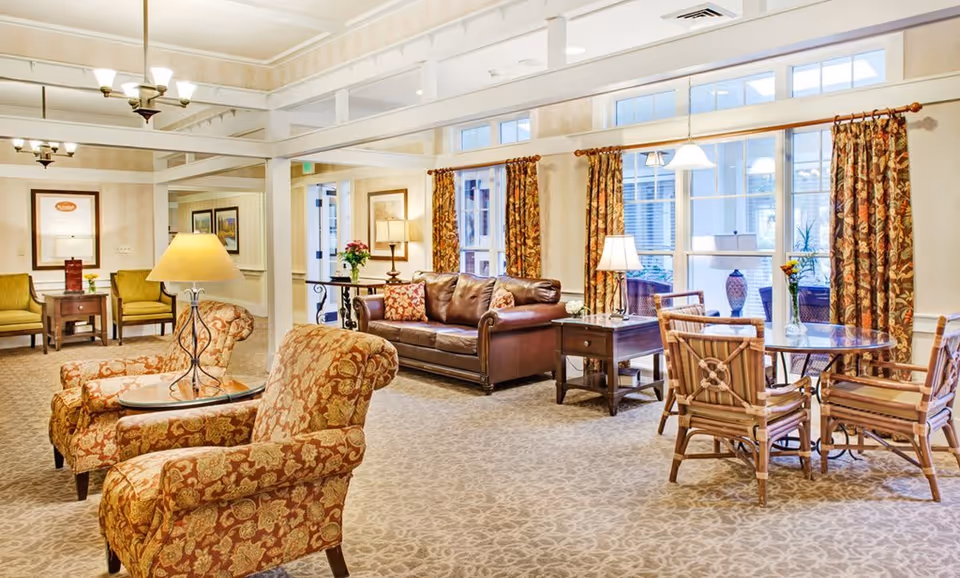 A bright and spacious living room area in a senior living facility with patterned armchairs, a brown leather sofa, wooden side tables with lamps, a round table with four chairs, large windows with floral curtains, and decorative vases on the window sill.
