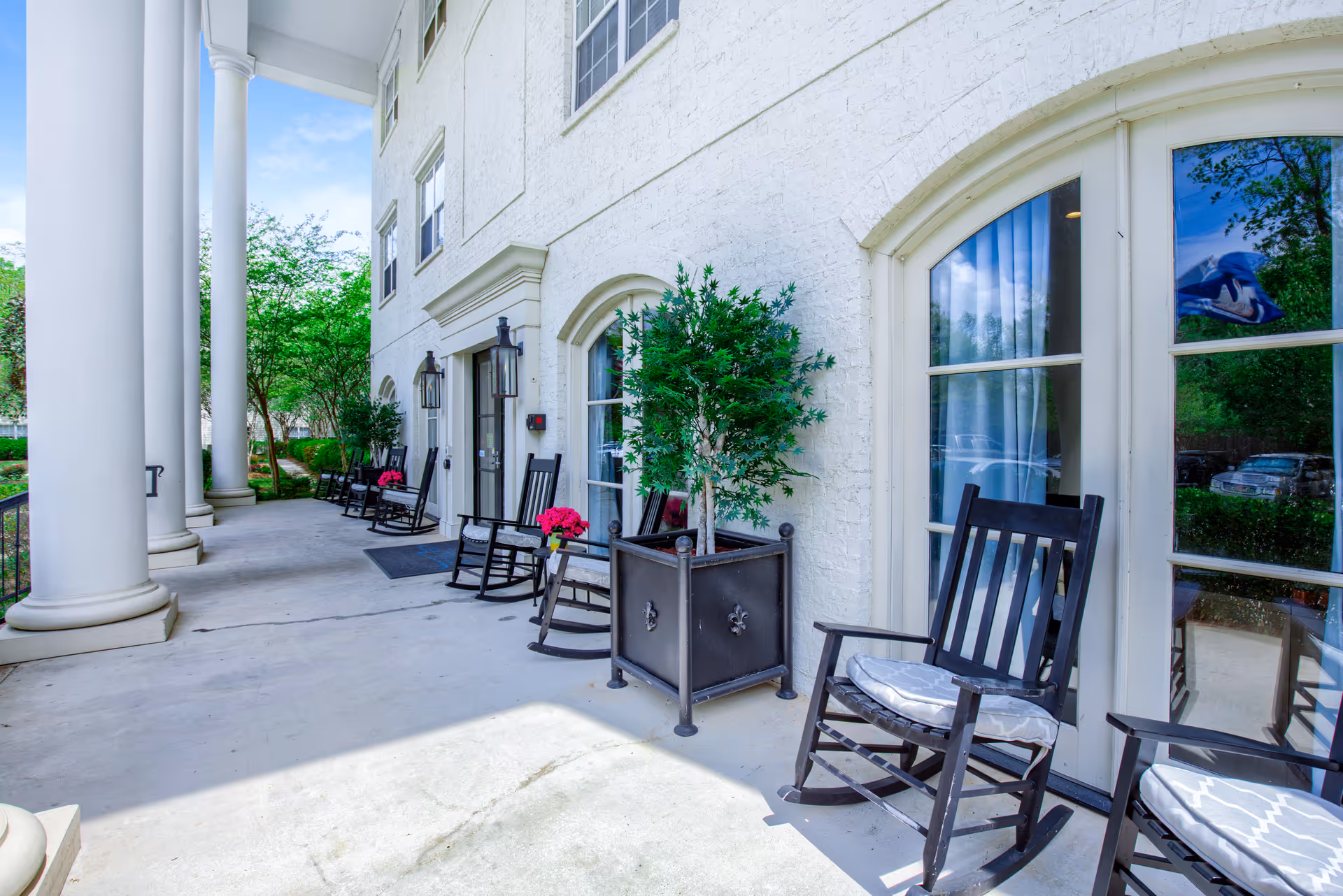 A covered outdoor patio area with large white columns, black rocking chairs with cushions, potted plants, and a white brick building with large windows and doors. Trees and greenery are visible in the background under a blue sky.