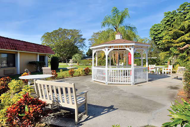Outdoor patio area with a white gazebo in the center, surrounded by benches and chairs. There are plants and bushes around the paved area, and a building with a red roof is visible on the left. The sky is clear and blue.
