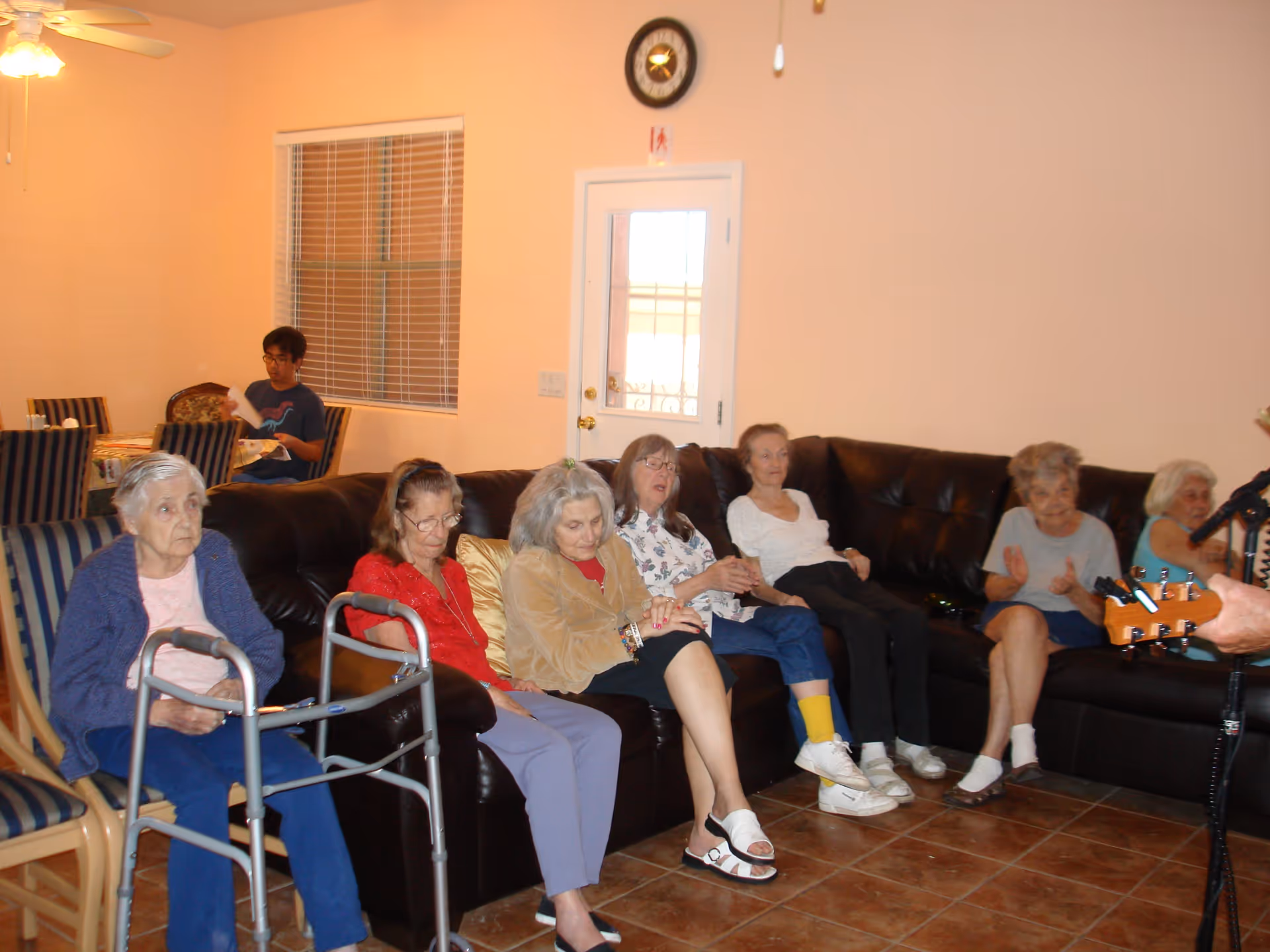 A group of elderly residents seated on a leather sectional in a living room while someone plays guitar.